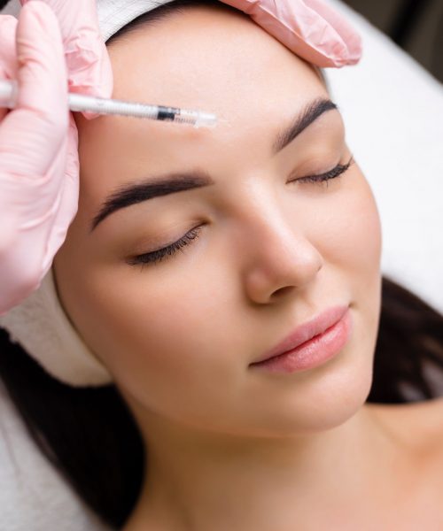 Close up of hands of young cosmetologist injecting botox in female face. She is standing and smiling. The woman is closed her eyes with relaxation.